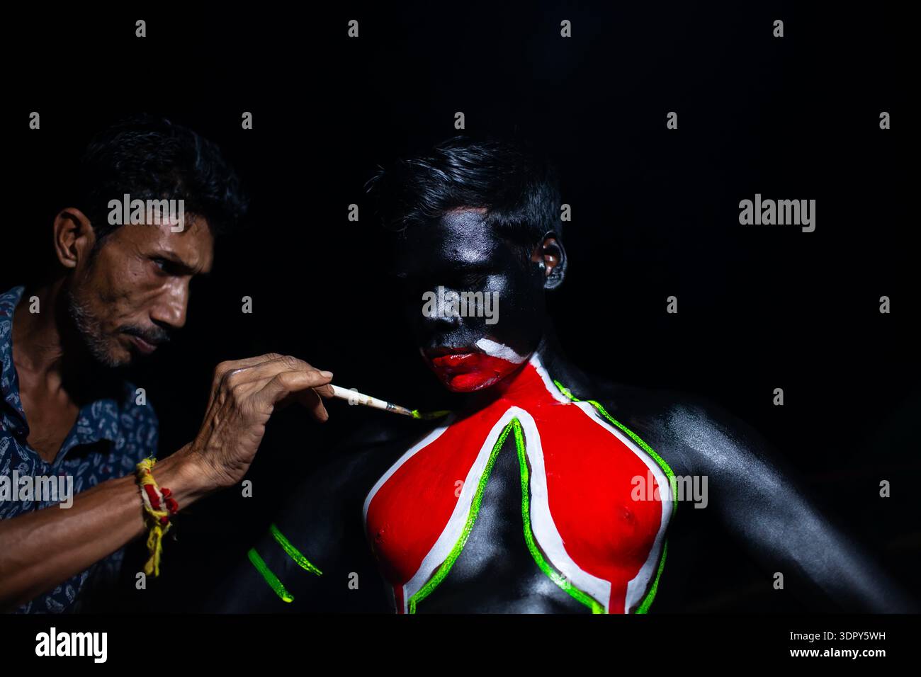 Sylhet, Bangladesch - 14. April 2025: Blick auf einen Mann, der akribisch leuchtende rote und grüne Designs auf die geschwärzte Haut eines anderen Mannes malte, wodurch eine Str. Stockfoto