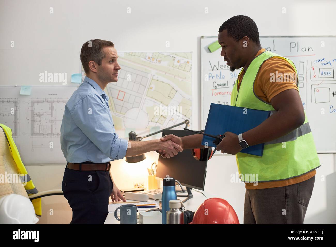 Kaukasischer Mann mittleren Alters, der am Schreibtisch steht und die Hände schüttelt, mit schwarzem Mann mittleren Alters, der Sicherheitsweste trägt, im Büro ein Klemmbrett hält, mit Bauplänen an der Wand Stockfoto