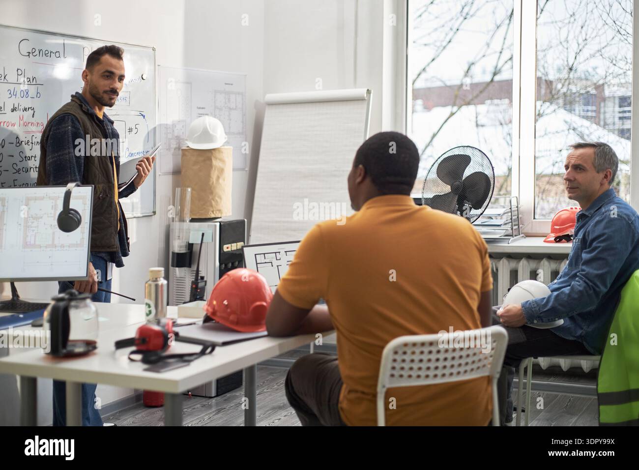 Kaukasier mittleren Alters, der neben dem Whiteboard steht und dem Schwarzen Mann mittleren Alters und dem Kaukasier mittleren Alters einen Projektplan vorstellt, der am Schreibtisch im Büro mit Baudokumenten sitzt Stockfoto