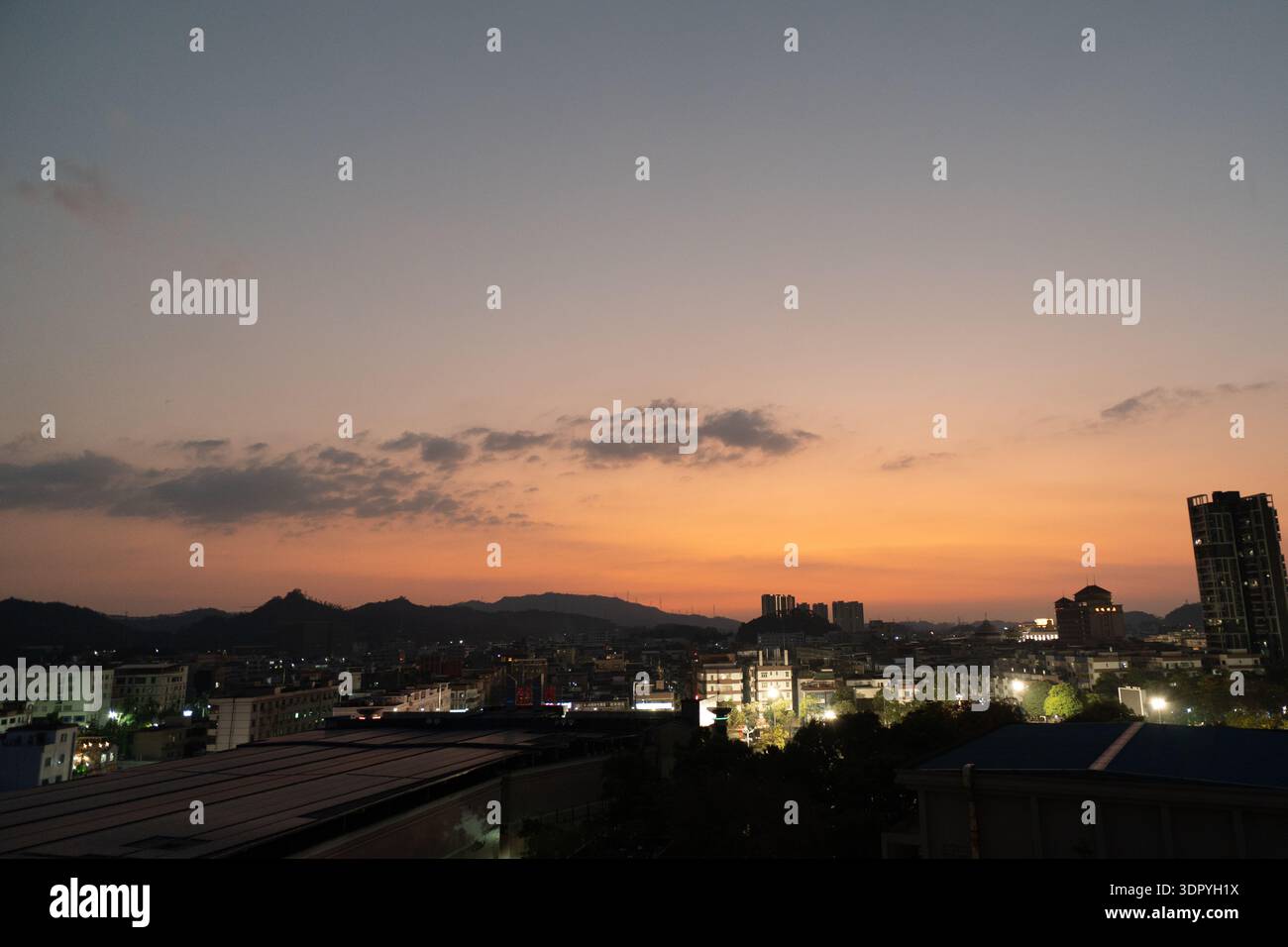 Blick auf den Sonnenuntergang über die Skyline der Stadt mit Bergen im Hintergrund, Stadtlichtern, die unter orangefarbenem und violettem Himmel leuchten Stockfoto