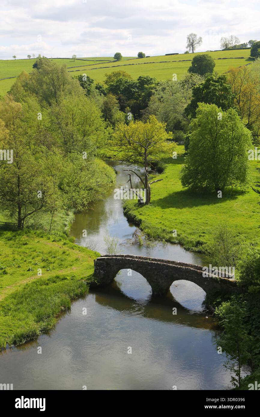 Ein erhöhter Blick auf eine alte Steinbrücke mit zwei Bögen, die einen ruhigen Fluss überspannt, während sie sich durch üppige grüne Wiesen und Felder im Peak schlängelt Stockfoto