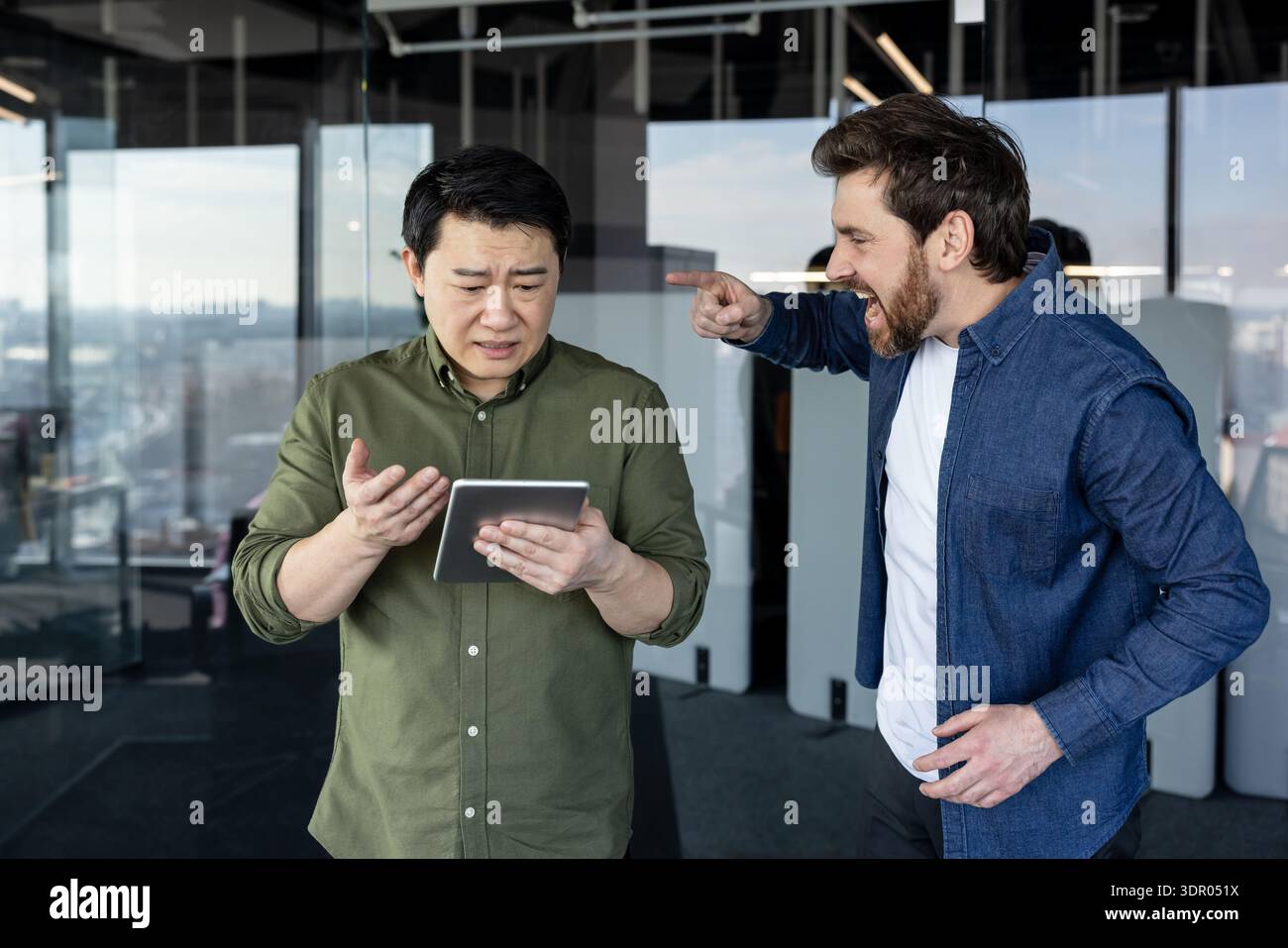 Ein wütender Geschäftsmann, der einen verwirrten asiatischen Kollegen mit einem digitalen Tablet anschreit und mit dem Finger auf ihn zeigt, der Konflikte am Arbeitsplatz, Belästigung und Stress in einer modernen Büroumgebung repräsentiert Stockfoto