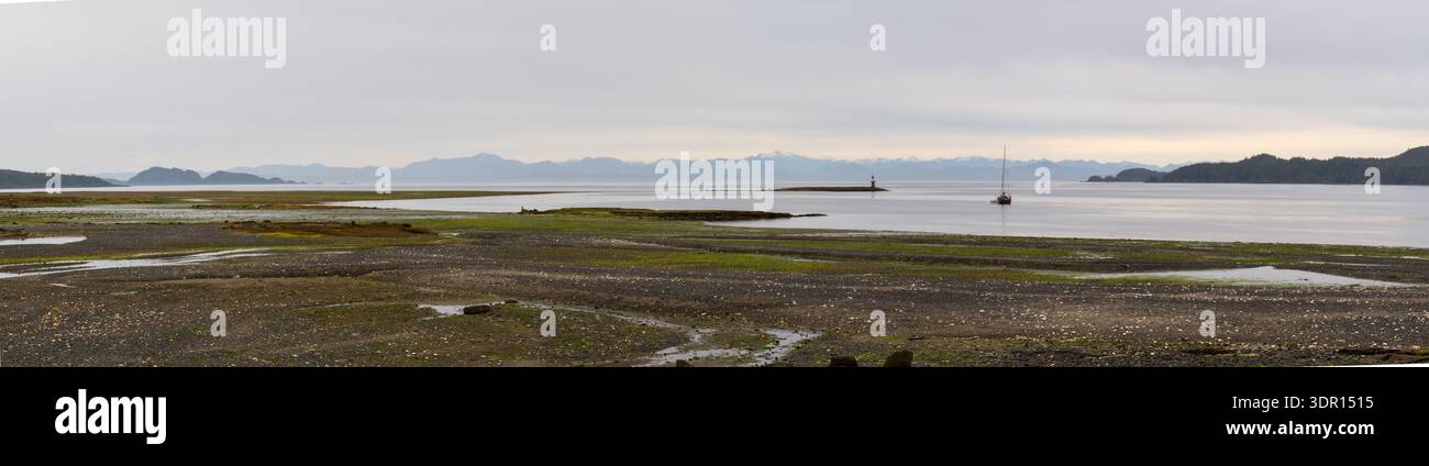 Port Hardy, BC, Kanada - 25. Juni 2024: Panoramablick auf ein Anker-Segelboot vor einem felsigen Strand von Port Hardy unter hohen Wolken und weit entfernten Bergen. Stockfoto