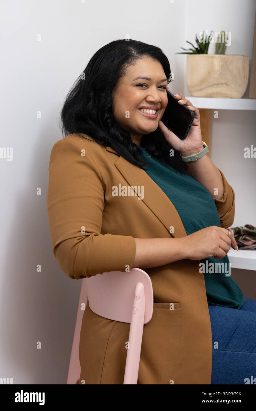 Frau in den 30ern, die auf rosa Stuhl sitzt, Smartphone am Ohr hält, in Blazer und Jeans lächelt Stockfoto