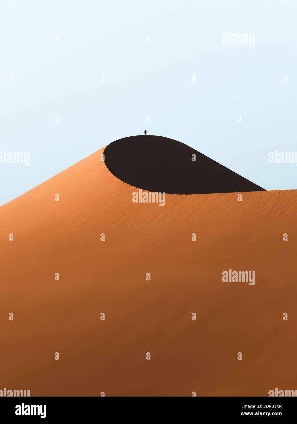 Der Blick auf eine majestätische orange Sanddüne wirft einen tiefen, umhüllenden Schatten unter einem hellen Himmel, eine einsame Figur auf der Düne, Hardap Region, Namibia. Stockfoto