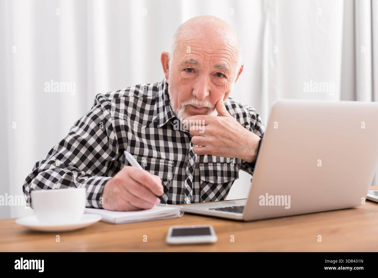 Ein älterer Mann sitzt an einem Tisch mit Laptop, Notizblock und Tasse Kaffee in einem hellen Raum Stockfoto