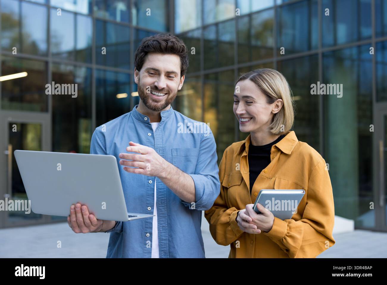 Geschäftskollegen stehen im Freien, ein Mann hält einen Laptop und gibt Gesten, während eine Frau mit einem Tablet lächelt, die Arbeit mit gemeinsamen Ideen und Teamarbeit diskutiert Stockfoto