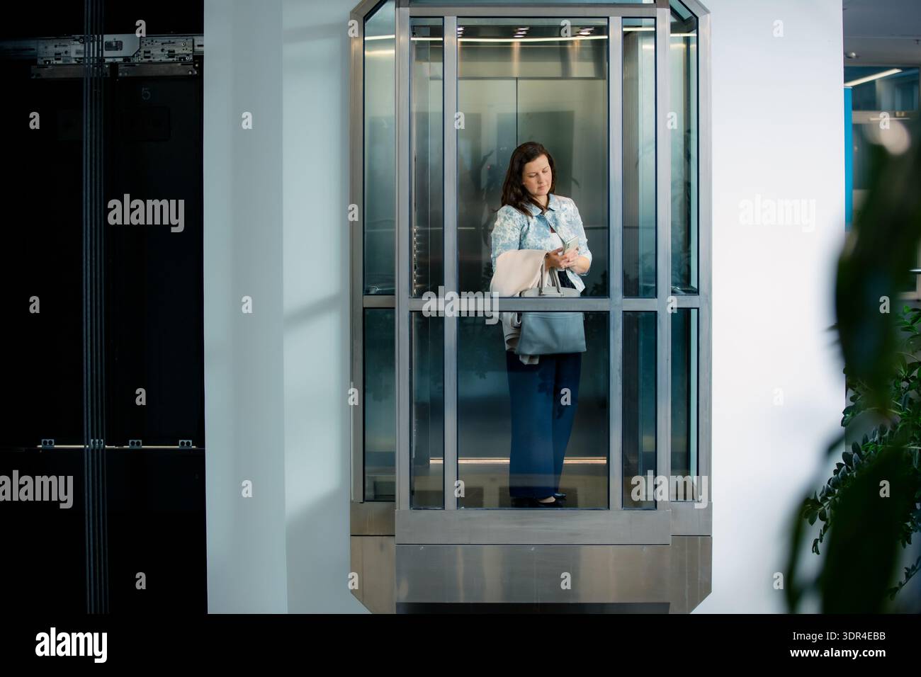 Professionelle Geschäftsfrau mit Smartphone im modernen Glasaufzug. Unternehmenskommunikation, Karrierefortschritt, Stockfoto