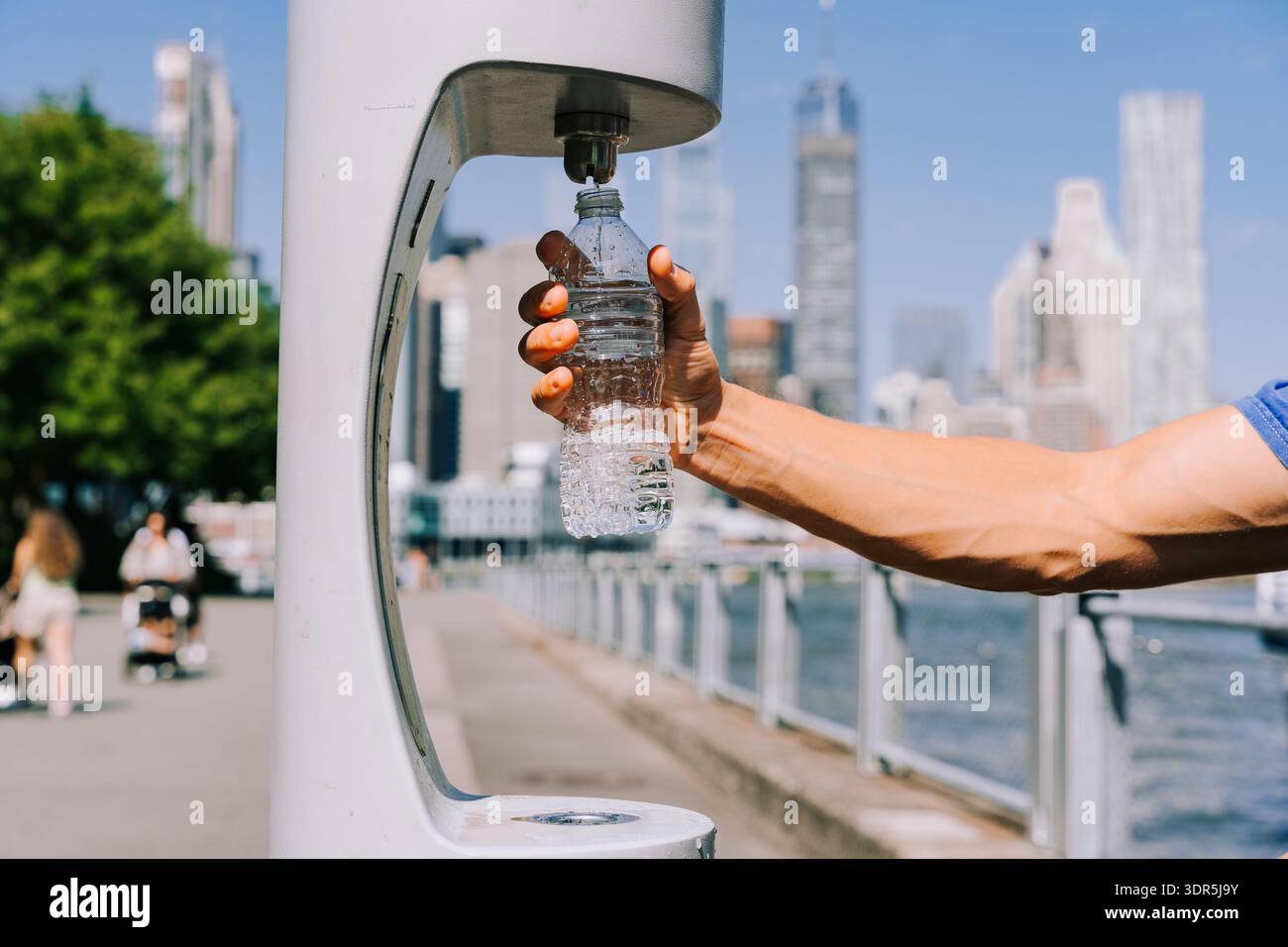 Eine Person füllt eine Wasserflasche am modernen öffentlichen Brunnen in der Nähe des Stadthafens auf und zeigt nachhaltige Trinkgewohnheiten und Bewegung in einem urbanen Außenbereich Stockfoto