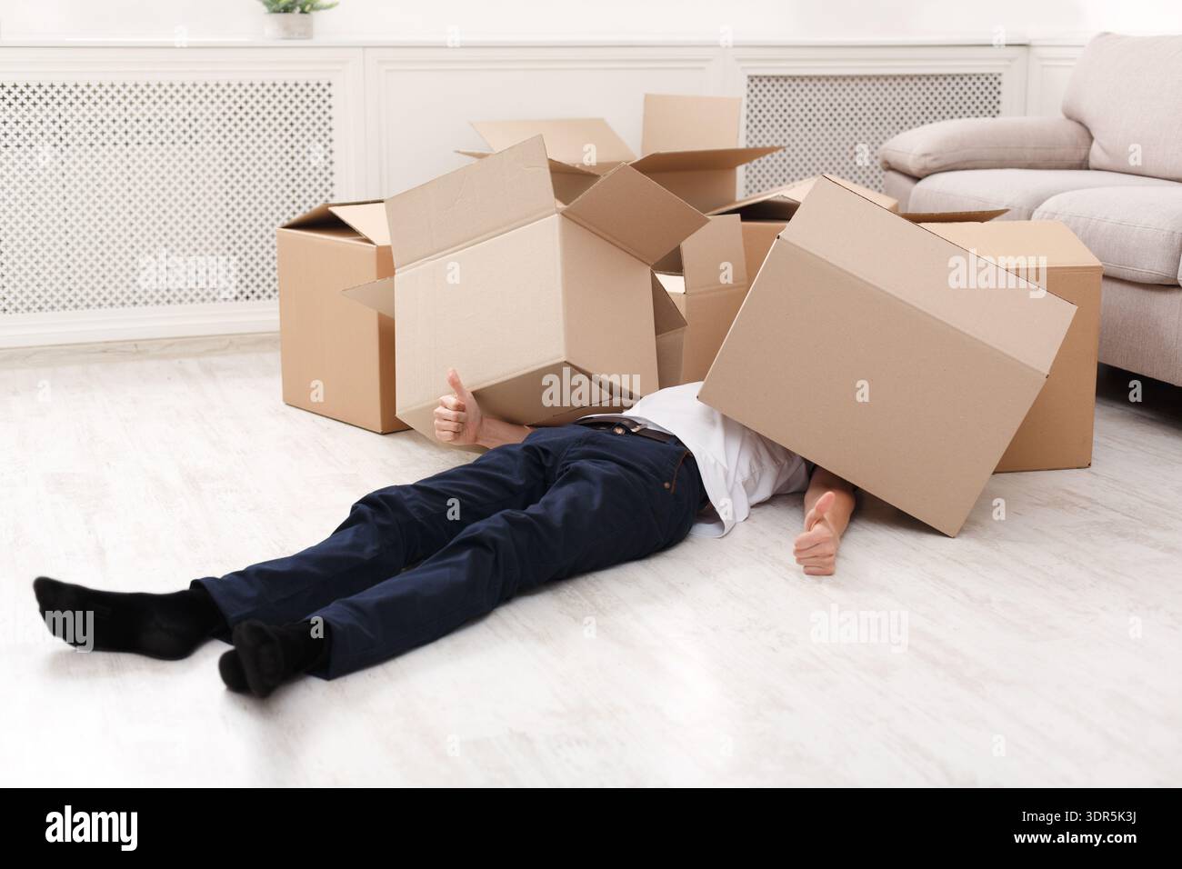Person Lying on Floor With Boxes Around and Box on Head in Living Room Setting Stockfoto