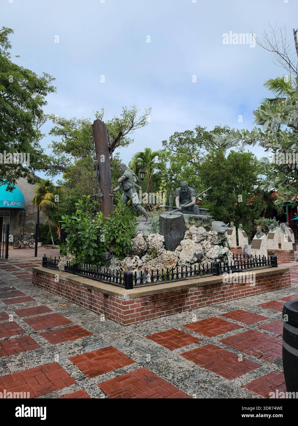 NUR FÜR REDAKTIONELLE ZWECKE - KEY WEST FLORIDA: Die Bronzeskulptur „Wreckers“ Memorial befindet sich im Key West Historic Memorial Sculpture Garden am Mallory Square. Das Denkmal zeigt historische Bergungsleute, die an einem Schiffswrack arbeiten, umgeben von üppigem tropischem Grün und Palmen unter hellem Himmel. Der gemauerte Innenhof und die Markise „Playhouse“ sind im Hintergrund zu sehen. - Smartphone-aufgenommenes Stockfoto