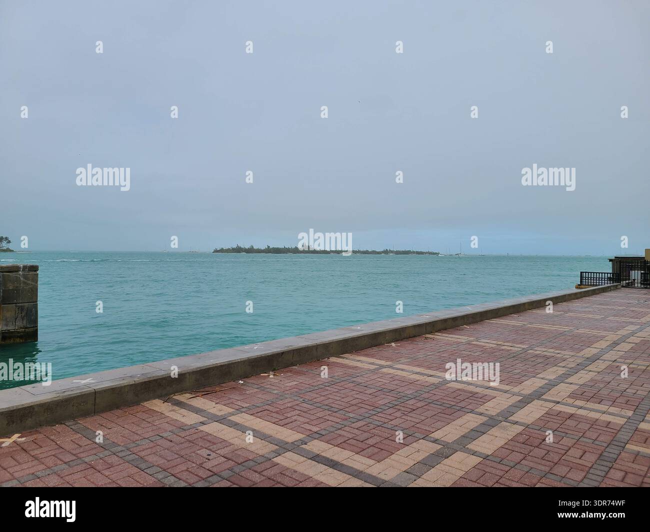 NUR FÜR REDAKTIONELLE ZWECKE - KEY WEST FLORIDA: Ein Hochwinkelblick mit Blick über das türkisfarbene Wasser von Key West Harbor in Richtung Sunset Key (Tank Island), Florida. Im Vordergrund befindet sich ein verwitterter Beton-Dock mit einem hellgelben Anlegepfosten und einer Sicherheitsleiter aus silbernem Metall. Die ferne Privatinsel ist gesäumt von üppigen Palmen und luxuriösen Küstenresidenzen unter einem bewölkten, stimmungsvollen tropischen Himmel. - Smartphone-aufgenommenes Stockfoto
