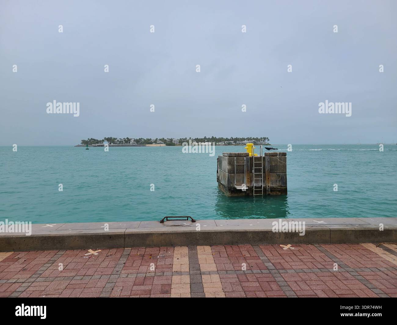 NUR REDAKTIONELLE VERWENDUNG - KEY WEST FLORIDA: : Ein Hochwinkelblick mit Blick über das türkisfarbene Wasser von Key West Harbor in Richtung Sunset Key (Tank Island), Florida. Ein Betondock mit einem gelben Anlegepfosten und einer Metallleiter befindet sich im Vordergrund, mit der üppigen, von Palmen gesäumten Privatinsel und Luxusresidenzen, die in der Ferne unter einem bewölkten, stimmungsvollen Himmel zu sehen sind. - Smartphone-aufgenommenes Stockfoto