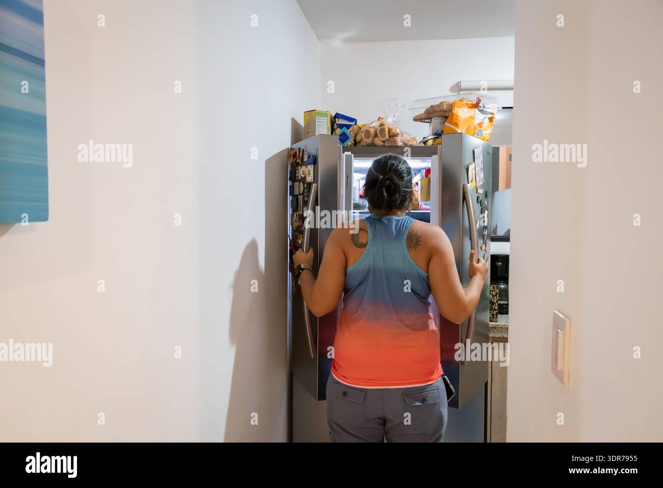Frau, die einen Kühlschrank öffnet und nach Essen in einer Küche sucht, nach einem Moment des privaten Lebensstils und dem täglichen Alltag drinnen. Stockfoto
