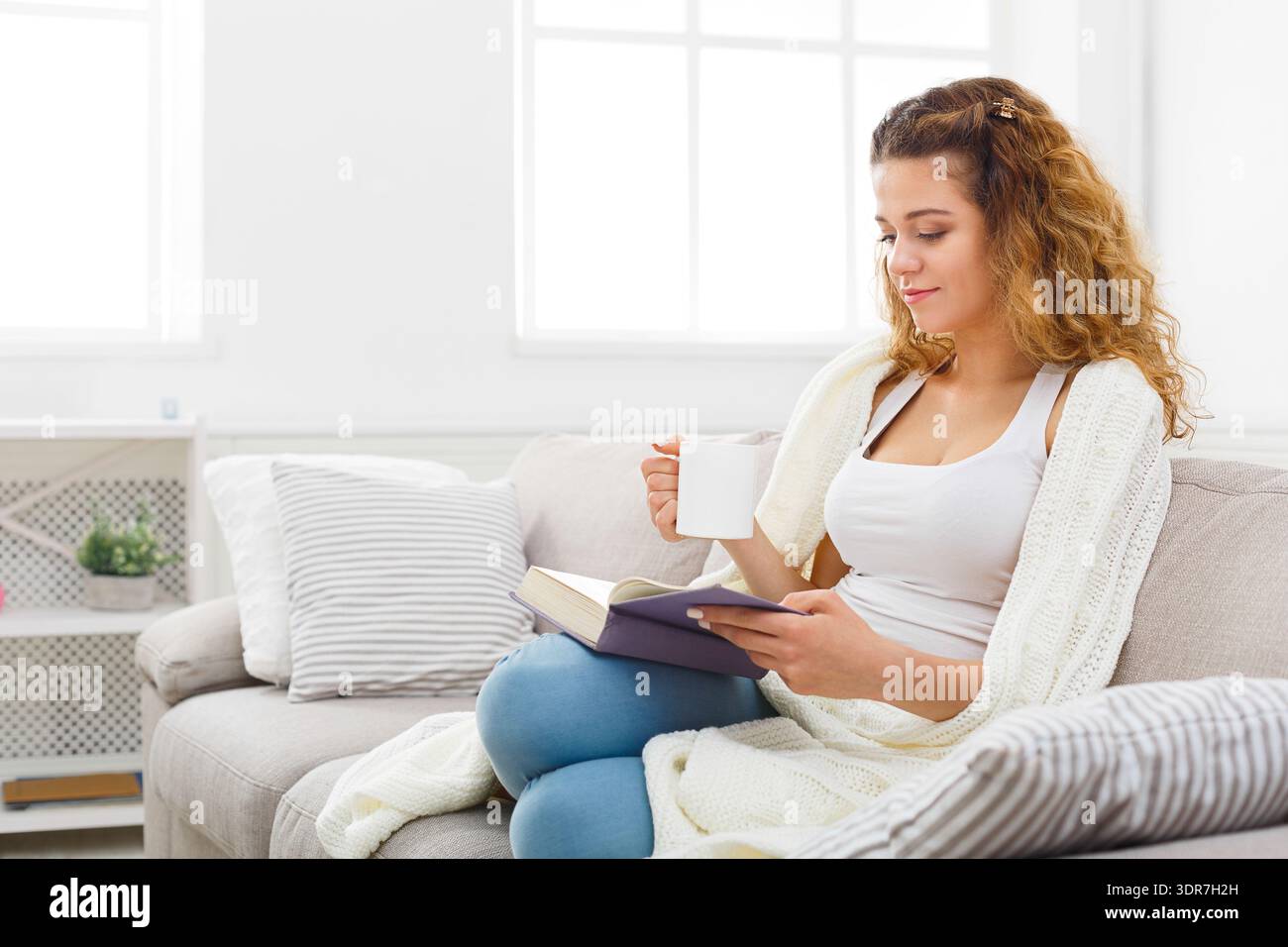 Frau, die auf einer Couch sitzt und ein Buch liest und während des Tages einen Becher in ihrem Haus hält Stockfoto