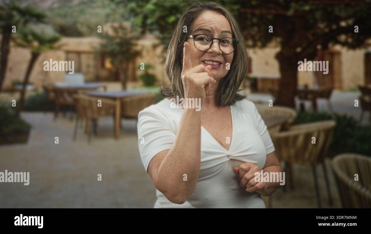 Hispanische Seniorin mit Brille zeigt mit dem Zeigefinger auf Terrassentische in einem Innenhof eines Cafés an einem Steingebäude; Vertrauensberatung. Stockfoto