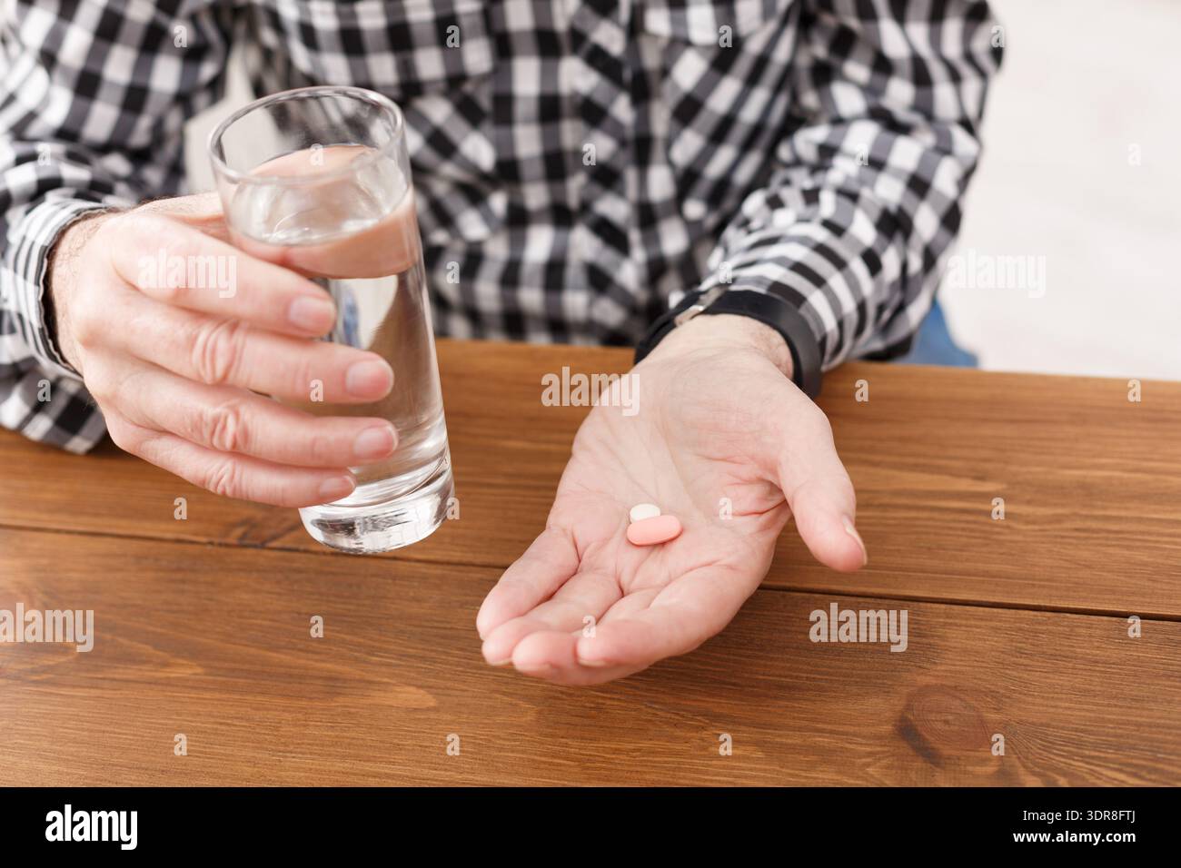 Person, die ein Glas Wasser und zwei Tabletten hält, während sie an einem Holztisch sitzt Stockfoto