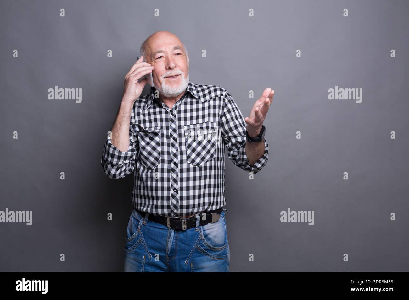Ein älterer Mann mit Bart hält ein Telefon am Ohr, während er ein kariertes Hemd und Jeans trägt Stockfoto