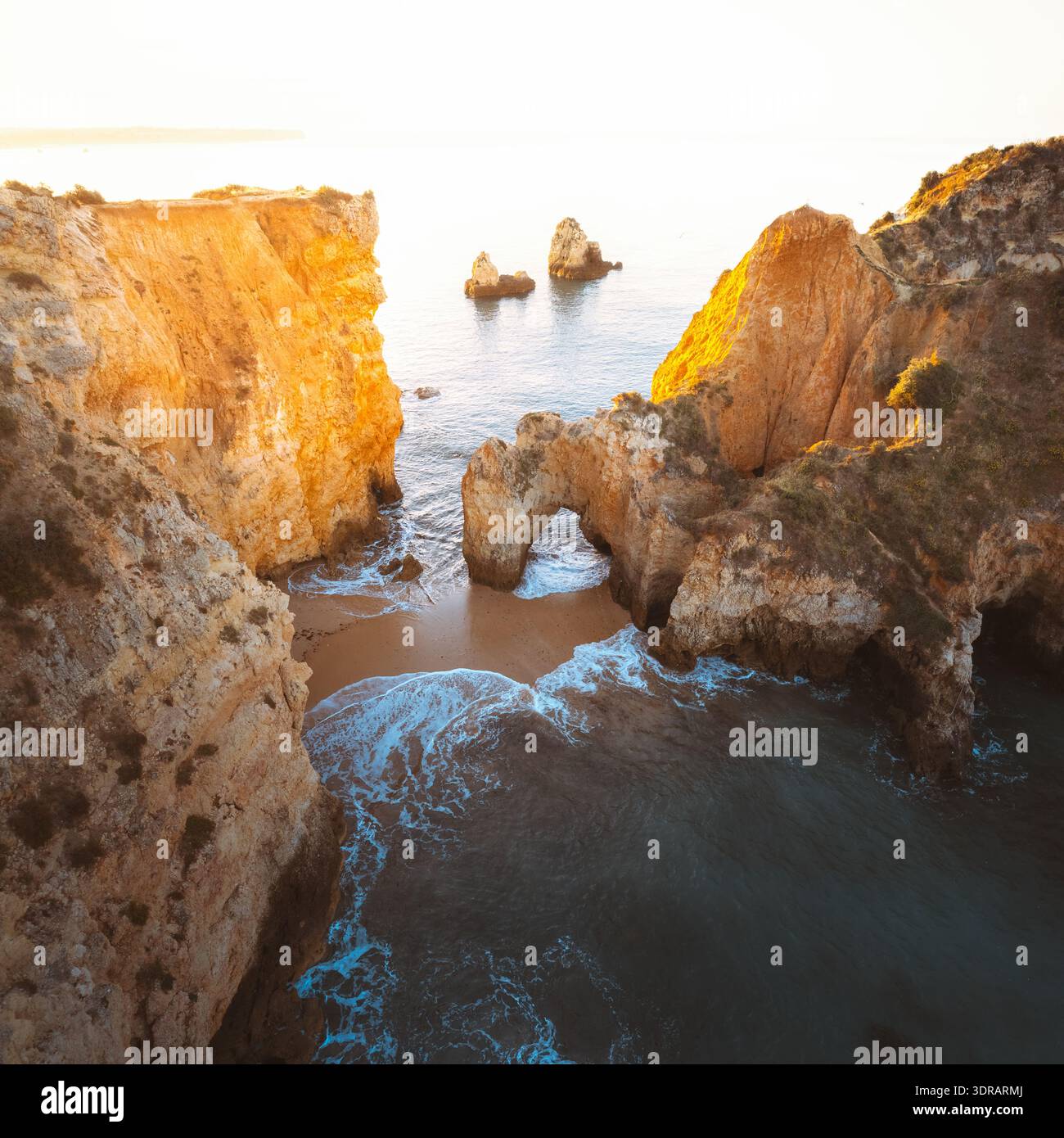 Aus der Vogelperspektive treffen auf dramatische Klippen auf das türkisfarbene Meer und bilden natürliche Bögen und abgeschiedene Strände unter dem warmen Glanz der Sonne, Praia da Marinha, F Stockfoto