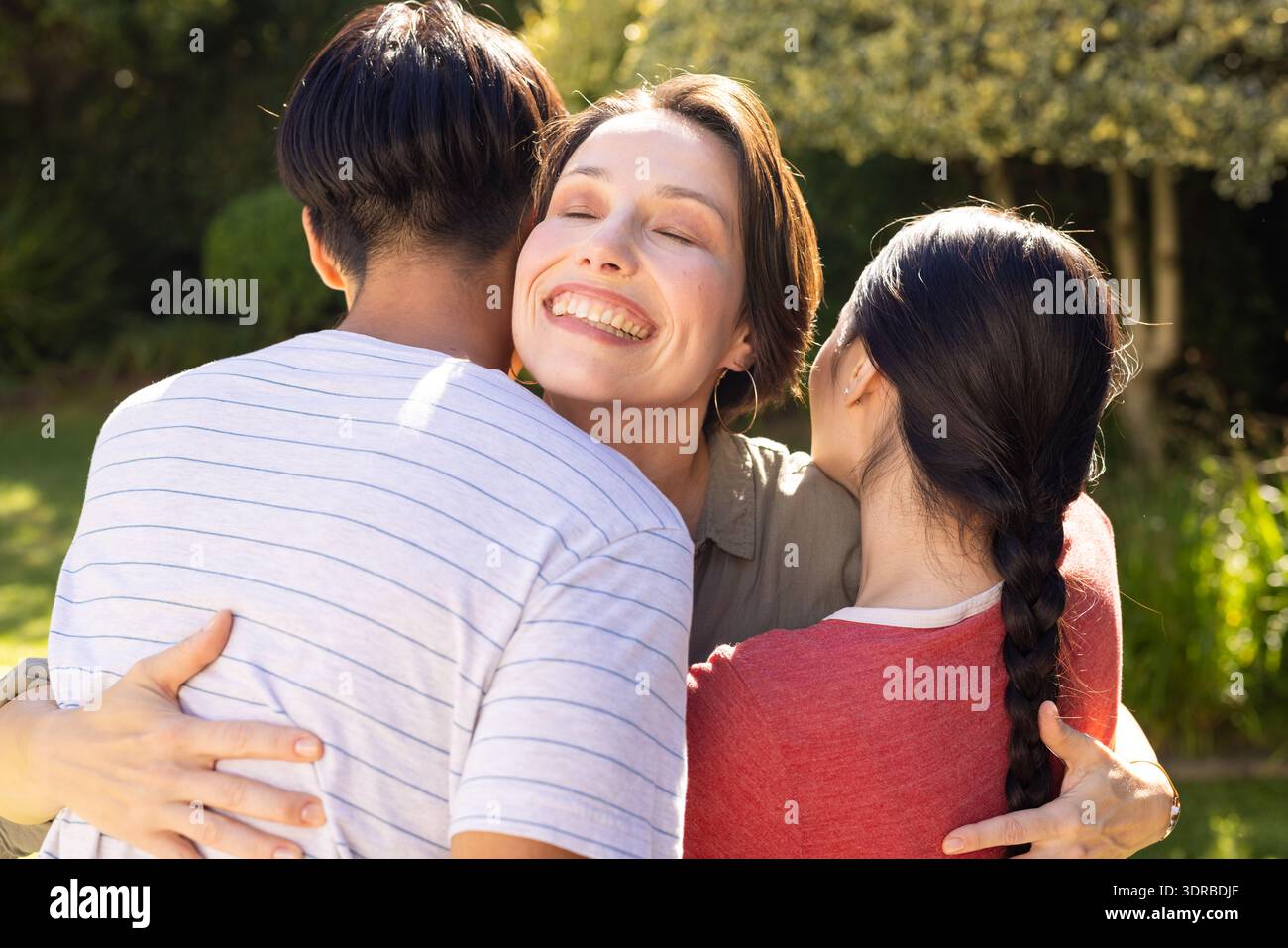 Lächelnde Frau umarmt Sohn und Tochter im Teenageralter und genießt die gemeinsame Familienzeit im Freien Stockfoto