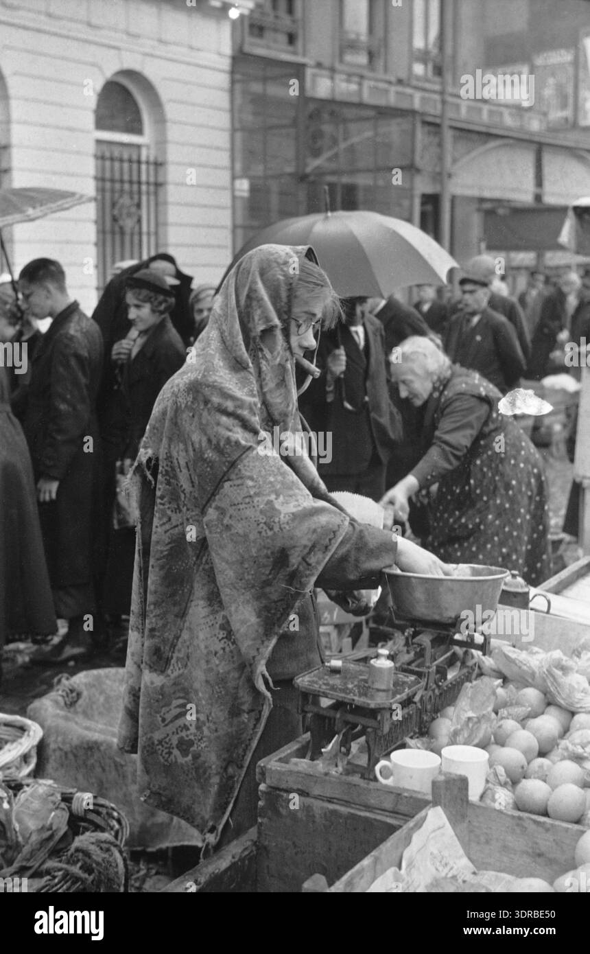 Das Foto zeigt einen Mann mit einer Zigarre im Mund, der bei regnerischem Wetter an einem Marktstand Orangen mit einer alten Tischdecke wiegt. Eine Reihe von Leuten mit Regenschirmen bewegt sich hinter ihm. Das Bild erfasst das niederländische Marktleben, den Straßenhandel, lokale Handelspraktiken, die Lieferantenaktivitäten und die sozialen Interaktionen auf einem Open-Air-Marktplatz und verdeutlicht die Marktbedingungen und das tägliche Leben in den Niederlanden im frühen 20. Jahrhundert. Stockfoto