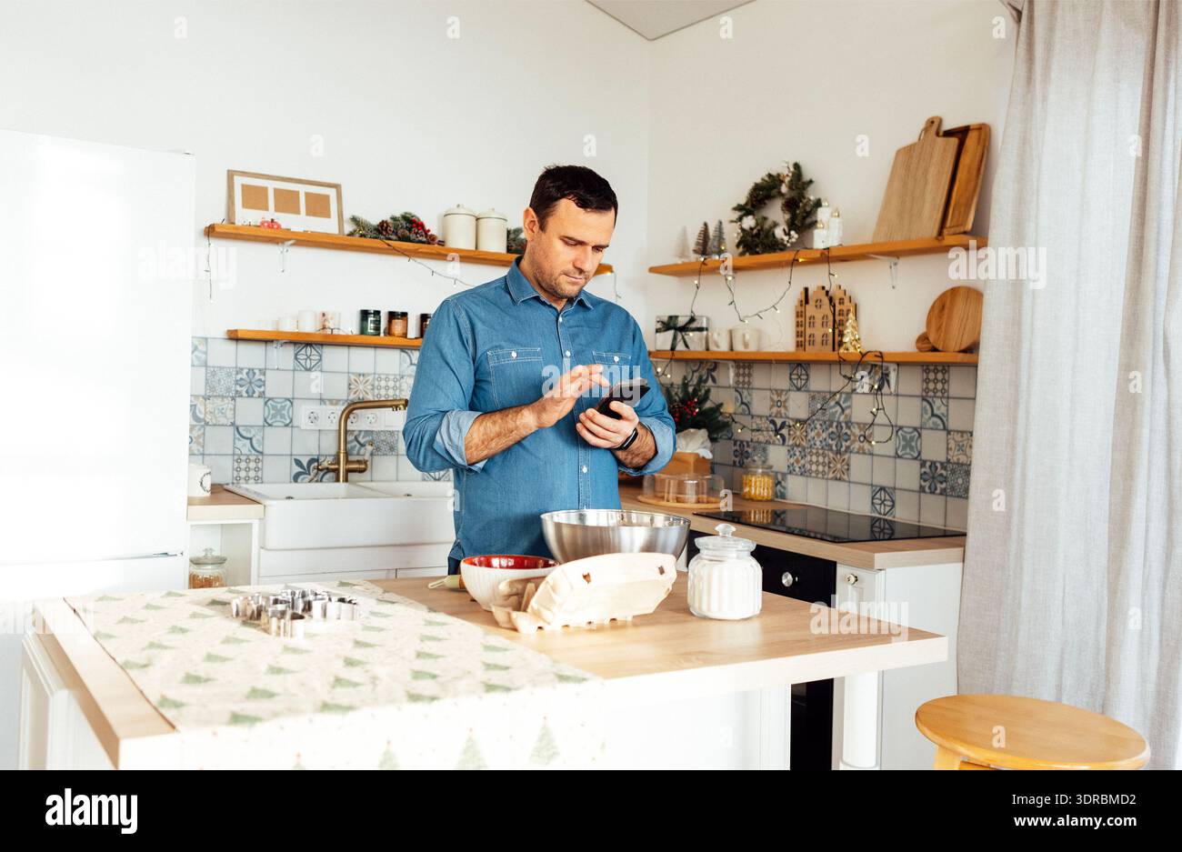 Ein attraktiver Mann in einem Jeansshirt kocht in der Küche zu Hause. Ein junger Mann hält ein Telefon und liest ein Rezept. Ein männlicher Mak Stockfoto