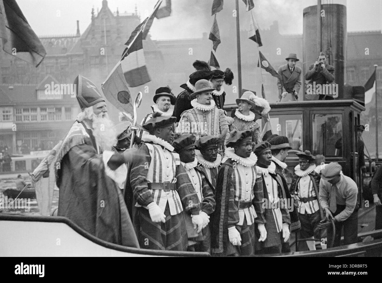 Dieses Bild zeigt Sint Nicolaas, dargestellt von Eduard Verkade, auf einem Dampfschiff, begleitet von Zwarte Pieten und spanischen Adligen während der Ankunft von Sinterklaas am Open Havenfront Amsterdam am 1. Dezember 1934. Centraal Station und Noord- en Zuid-hollandsch Koffiehuis sind im Hintergrund zu sehen. Einige Zwarte Pieten waren Suriname Crew-Mitglieder. Das Foto zeigt eine Seefahrerparade, traditionelle Trachten, öffentliche Zuschauerschaft und niederländische Feiertage, die vom Initiatief Comité Amsterdam organisiert werden. Stockfoto