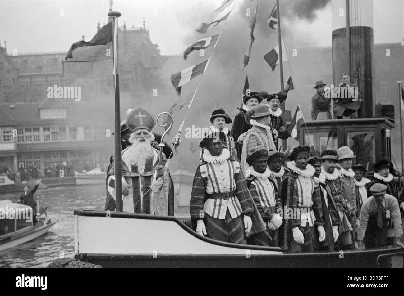 Das Foto zeigt Sint Nicolaas, unterstützt von Eduard Verkade, auf einem Dampfschiff mit einem stark rauchenden Trichter, begleitet von Zwarte Pieten und spanischen Adligen während der Ankunft der Sinterklaas am Open Havenfront Amsterdam am 1. Dezember 1934. Im Hintergrund erscheinen die Centraal Station und Noord-Zuid Hollandsch Koffiehuis. Das Bild zeigt festliche Seeverkehrsprozession, öffentliche Zuschauer, zeremonielle Kostüme, städtische Kanäle und niederländische Feiertage, die vom Initiatief Comité Amsterdam organisiert werden. Stockfoto