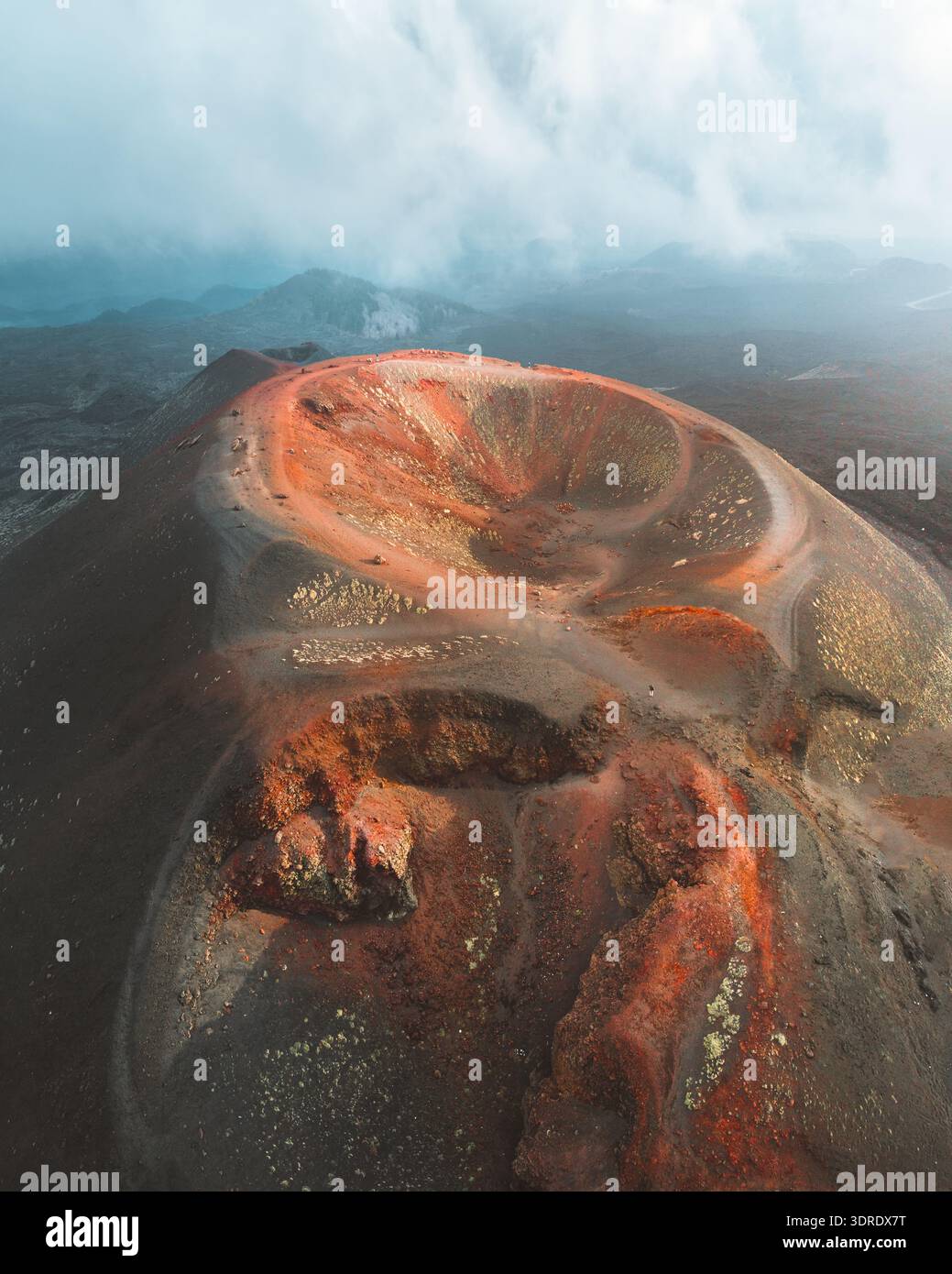 Aus der Vogelperspektive der vulkanischen Landschaft, wo das dunkle, zerklüftete Gelände auf den leuchtenden roten Krater trifft, geküsst von der weichen Berührung der Wolken, Nicolosi, Sicil Stockfoto