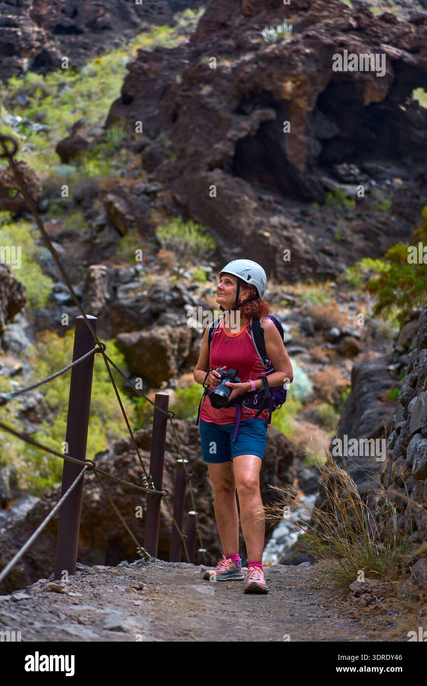 Weibliche Wanderer mit Kamera und Helm, die auf dem Masca Gorge Trail mit vulkanischen Felswänden dahinter aufblickt Stockfoto