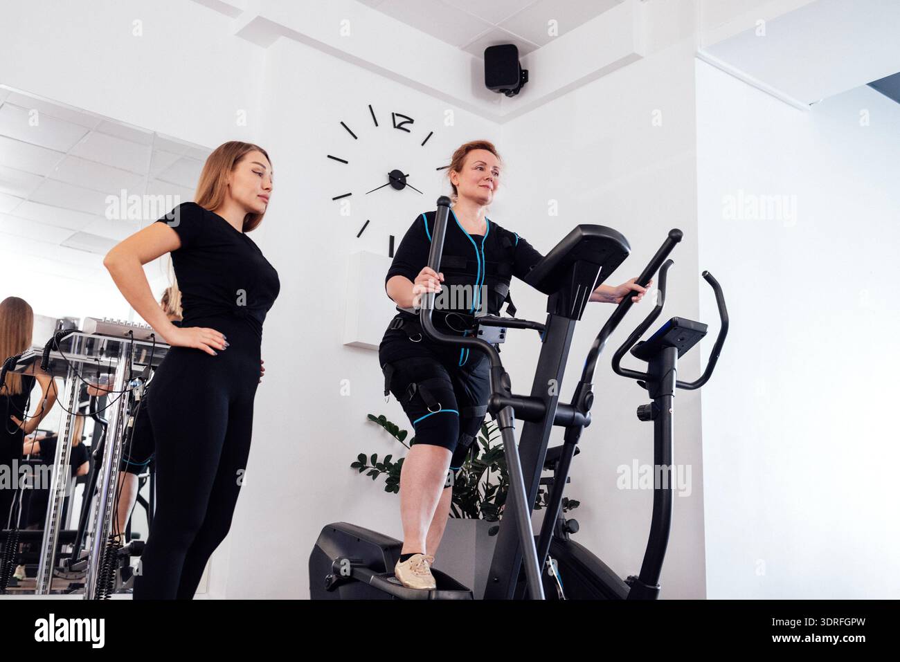 Ältere Frau im Anzug für elektrische Muskelstimulation trainiert auf einem Ellipsentrainer mit ihrem persönlichen Trainer im Fitnessstudio oder Fitness Stockfoto