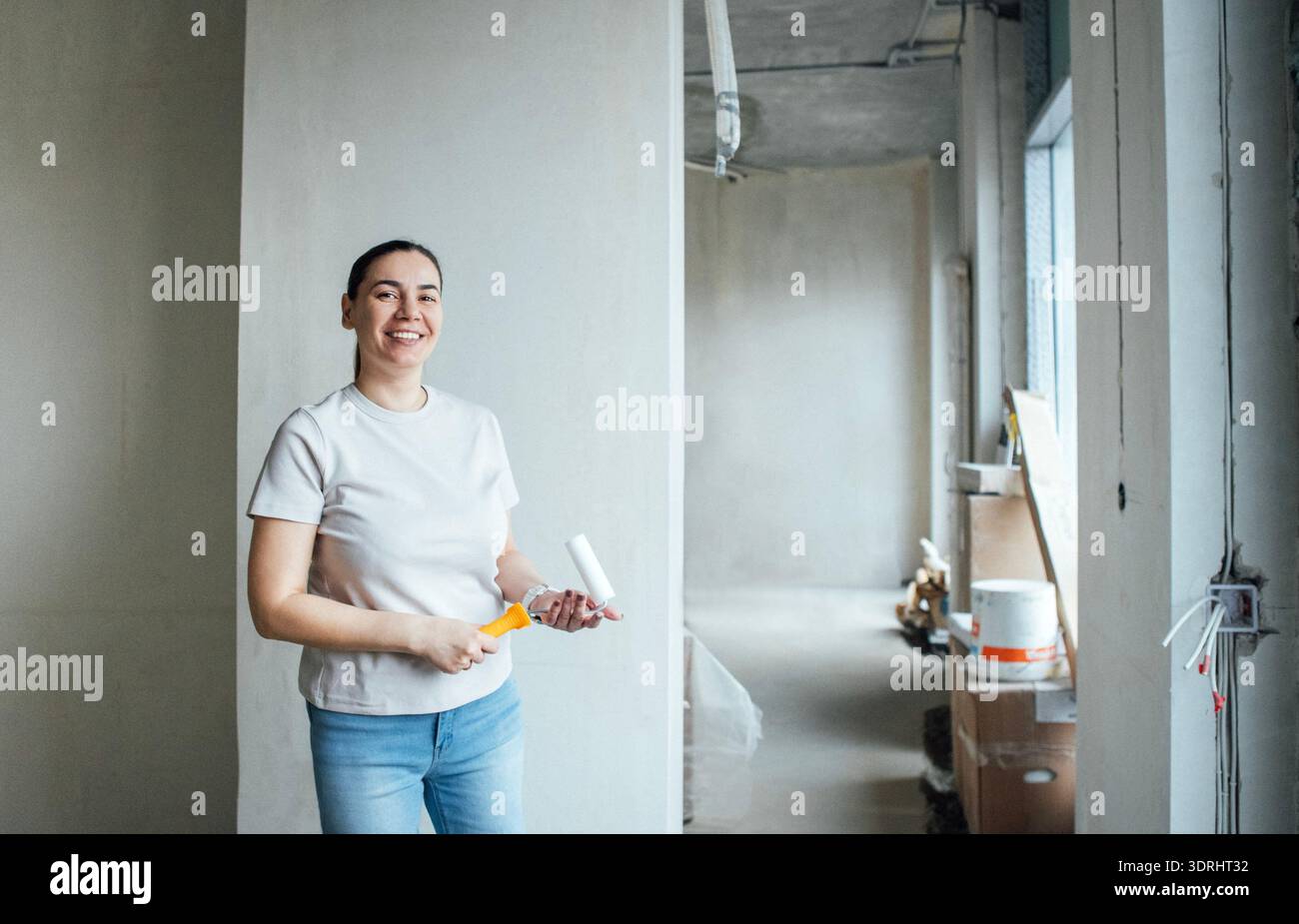 Eine Frau mittleren Alters hält eine Malerwalze und lächelt. Eine junge Dame steht in einem hellen, teilweise renovierten Zimmer mit unvollendetem W Stockfoto