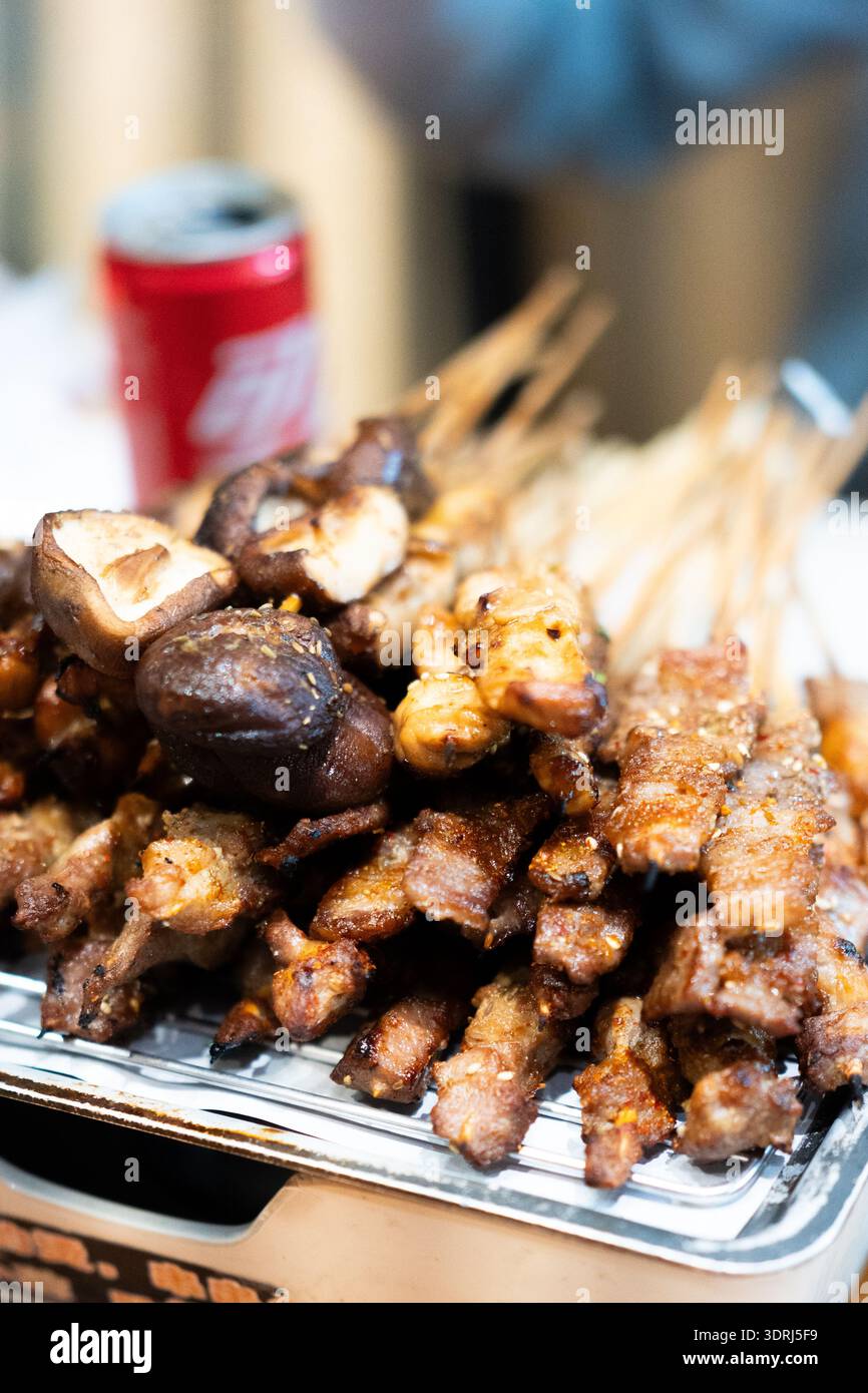 Chinesische Streetfood-Spieße mit gegrilltem Fleisch und Pilzen auf Metalltablett, rote Dose im Hintergrund Stockfoto