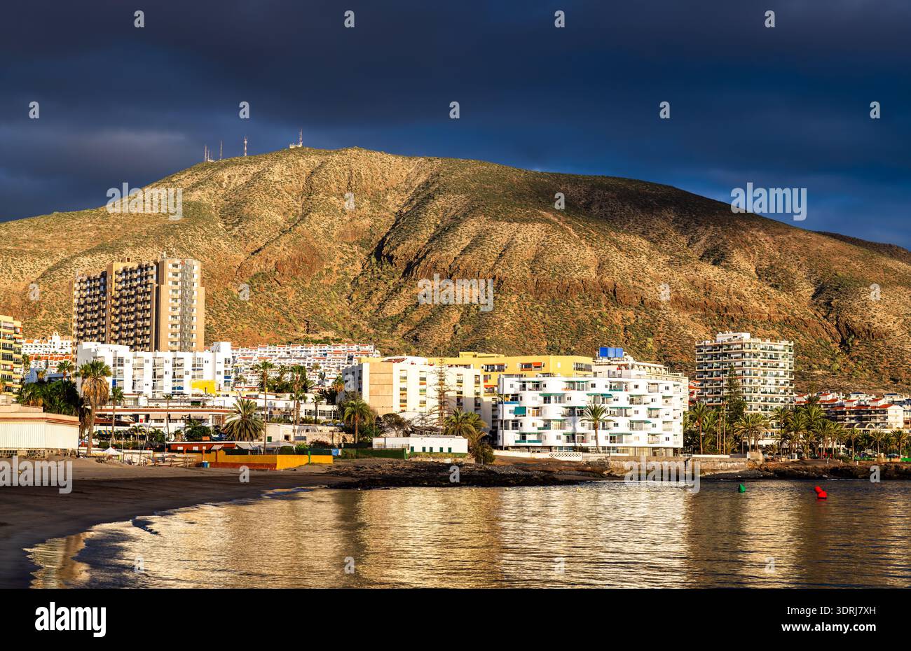 Blick auf den Strand Playa de Los Cristianos und den Berg Guaza auf Teneriffa, Kanarischen Inseln, Spanien. Mit Sandstrand, ruhigem Meereswasser und bei Sonnenuntergang beleuchteten Resortgebäuden. Stockfoto