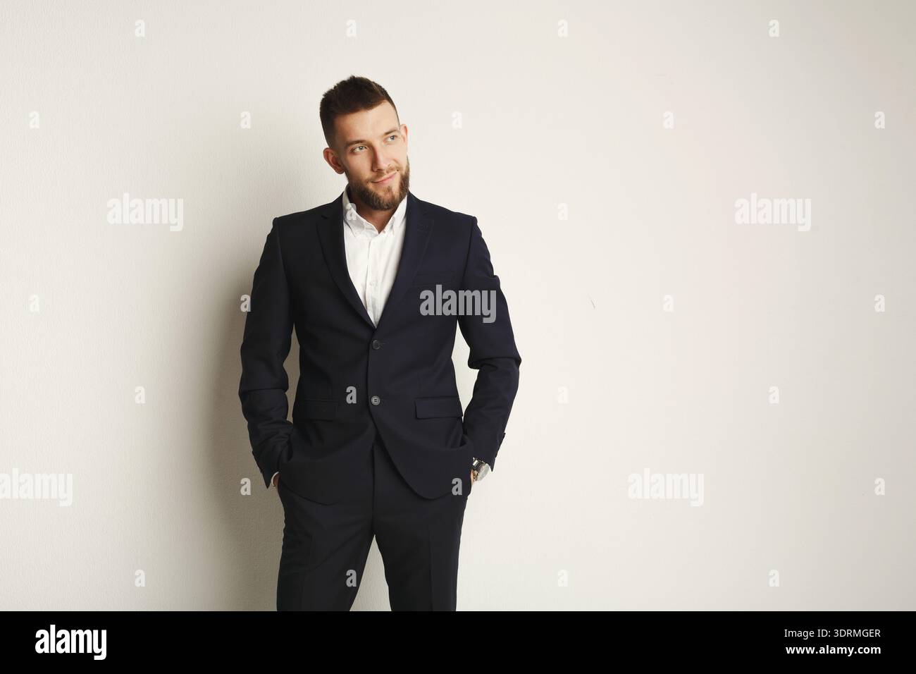 Mann in einem Anzug, der tagsüber in einem Studio an einer Plain Wall steht, mit Händen in Taschen Stockfoto