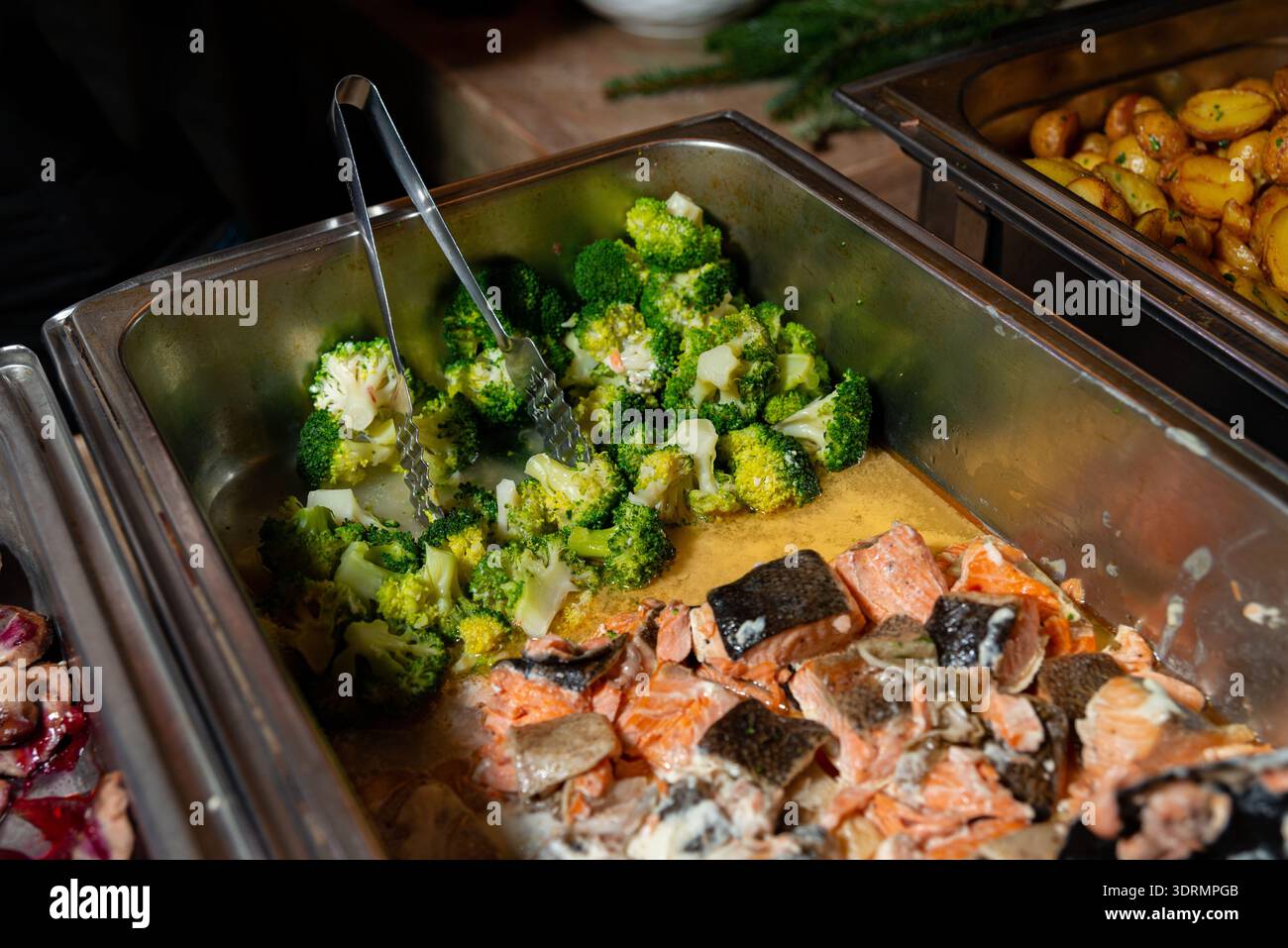 In der Edelstahl-Pfanne werden gedämpfte Brokkoli-Röschen sowie gekochte Lachsstücke mit Haut serviert, wobei eine Metallzange in der Pfanne liegt Stockfoto