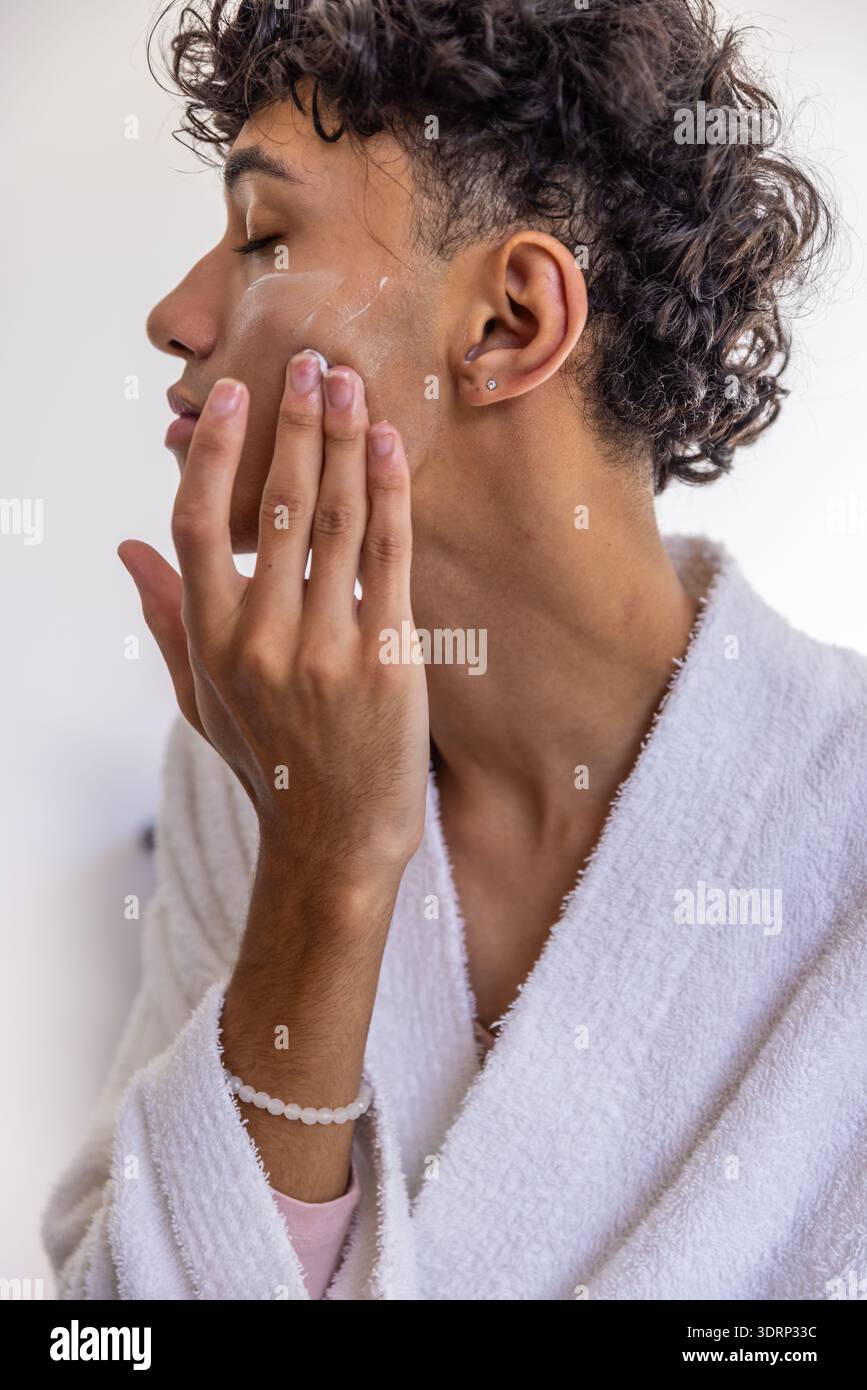 Mann in weißem Bademantel, der Gesichtscreme auf die Wange im Badezimmer aufträgt und Armband und Ohrstecker trägt. Hautpflege, Selbstpflege, Wellness Stockfoto