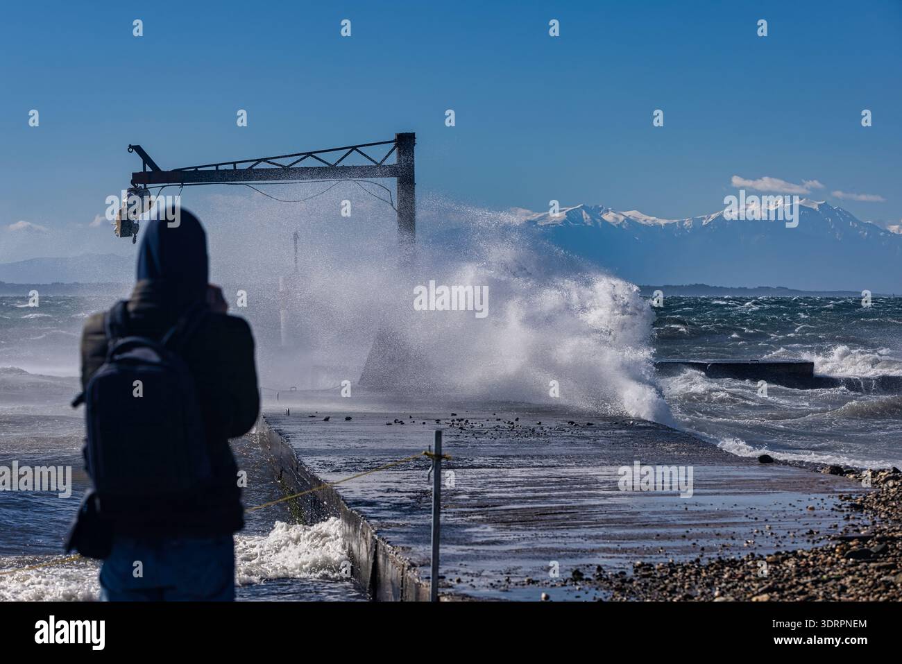 Eine Person steht auf einem Pier an der Küste, während mächtige Wellen gegen die Struktur prallen und Wasser in die Luft schicken. Ein Metallkran erhebt sich über die Szene Stockfoto