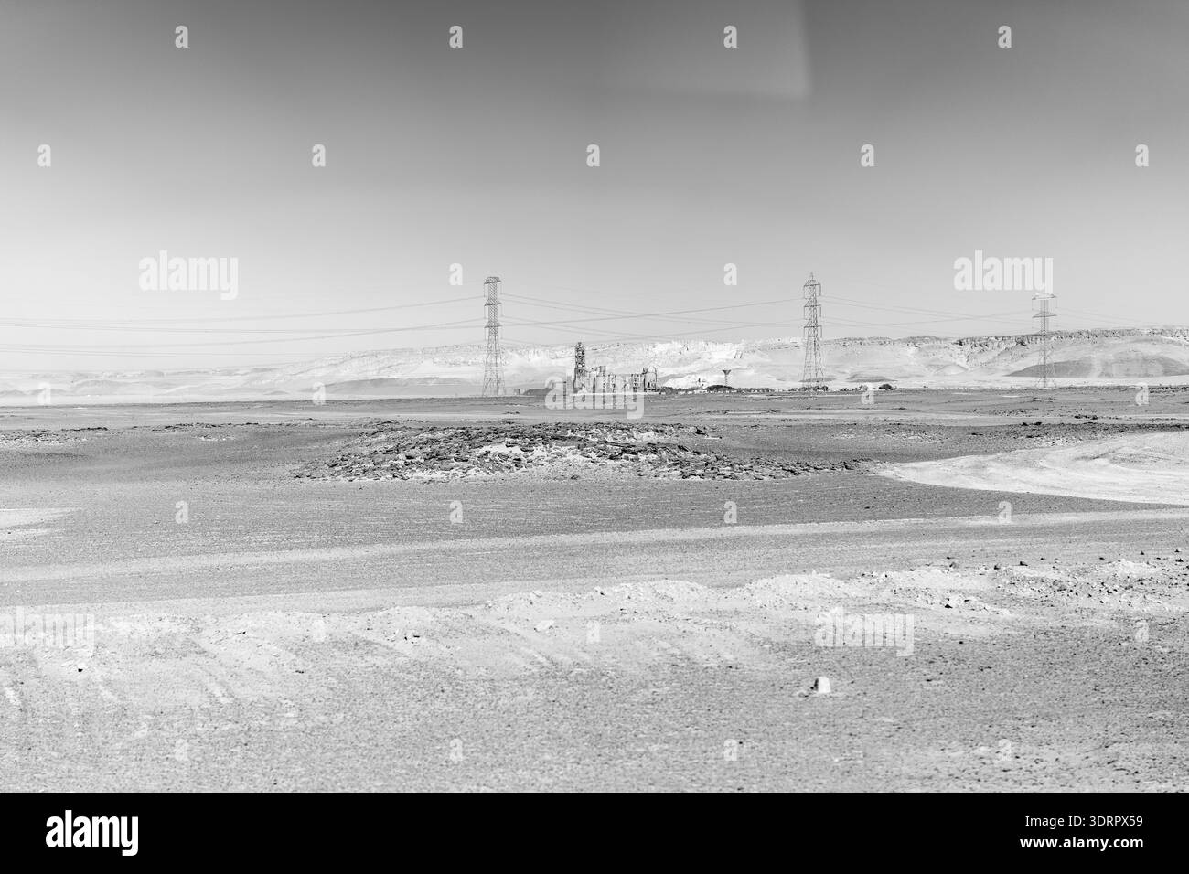 Schwarzweiß-Ansicht einer rauen Wüste in der Nähe von Assuan mit Stromleitungen und Industriebauten, die Isolation, Größe und Umweltauswirkungen vermitteln. Stockfoto