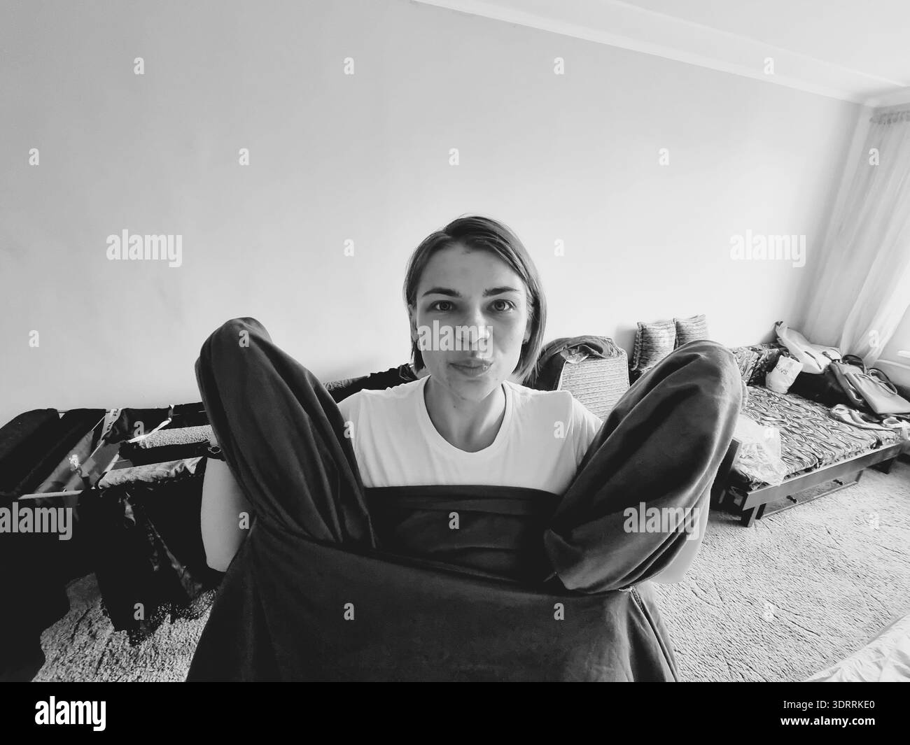 Junge Frau, die lässig in einem gemütlichen Wohnzimmer sitzt, halb bedeckt von dunkler Kleidung, lächelt und in einem ehrlichen Schwarz-weiß-Leben in die Kamera blickt Stockfoto