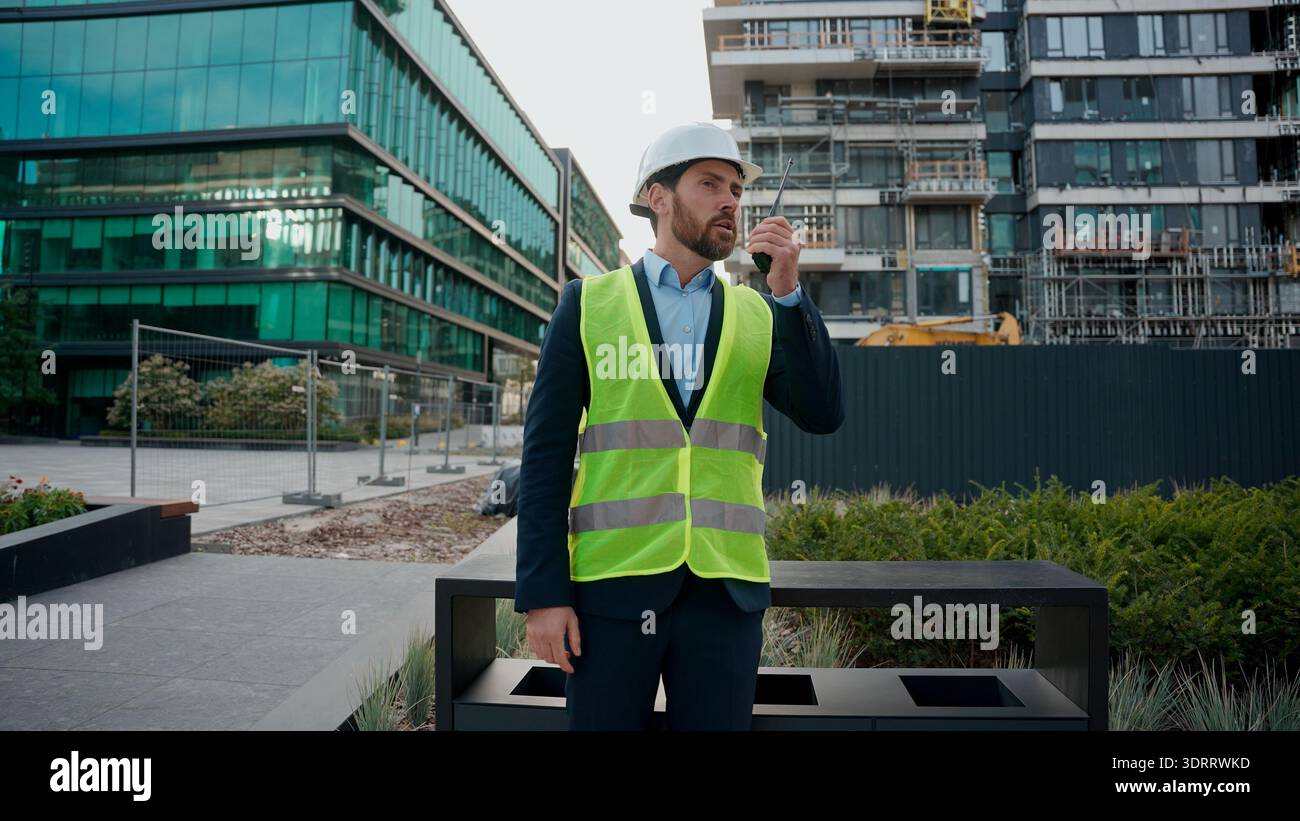 Moving Shot Kaukasischer Mann Bauingenieur in Hardhat Talk Remote Radio Outdoor Urban Building Architekt Bauunternehmer mit Helm Sicherheitsweste Stockfoto