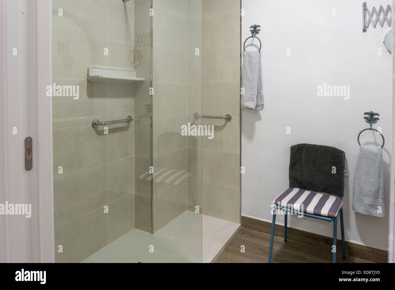 Barrierefreie, ebenerdige Dusche mit Glastrennwand, Haltegriffen, Regal und hellen Fliesen, die das Altern fördern Stockfoto