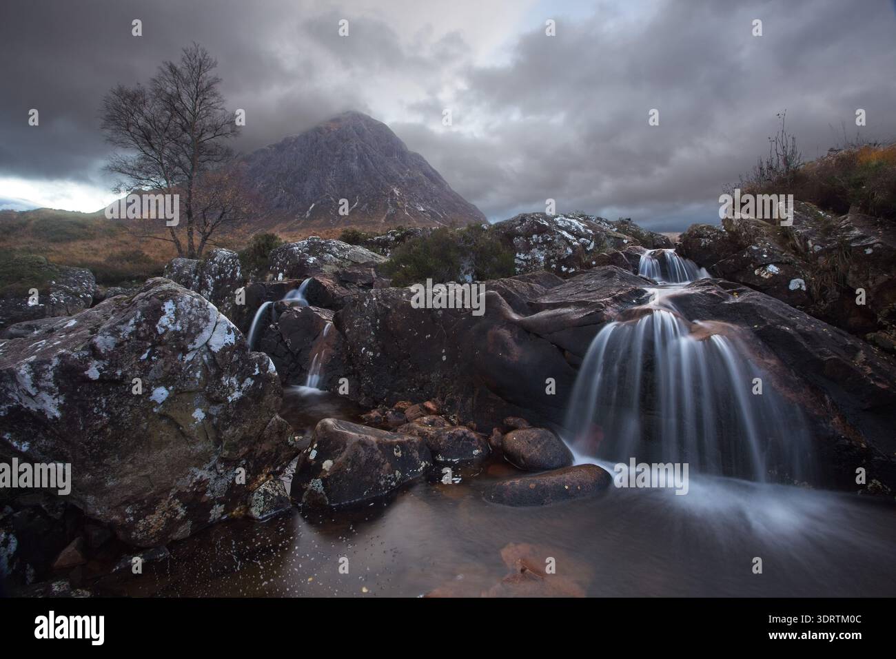 Kleine Wasserfälle, die über dunkle Felsen mit Bergen und stürmischem Himmel in den schottischen Highlands, Glen Etive, fließen Stockfoto
