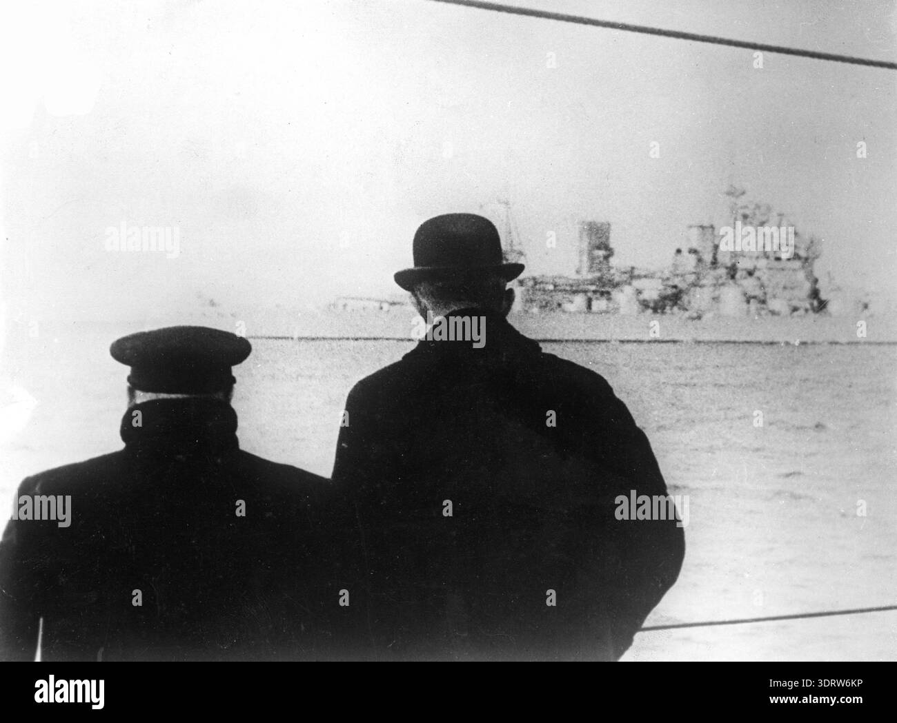 Dieses Schwarzweiß-Foto aus dem Jahr 1941 zeigt Winston Churchill und den britischen Botschafter Lord Edward Halifax von hinten, die am Geländer eines kleinen Dampfers stehen. Im Hintergrund ist das gewaltige Kriegsschiff HMS King George V. sichtbar, was ein Gefühl von Kriegsbedeutung und drohender Reise schafft. Stockfoto