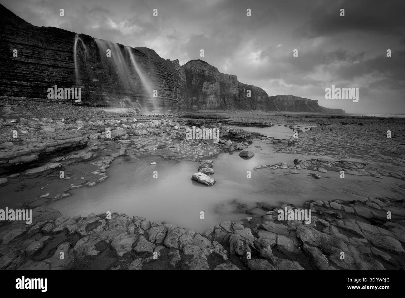 Schwarz-weiß lange Exposition eines Wasserfalls, der von Klippen auf ein felsiges Ufer entlang der walisischen Heritage Coast fällt. Stockfoto