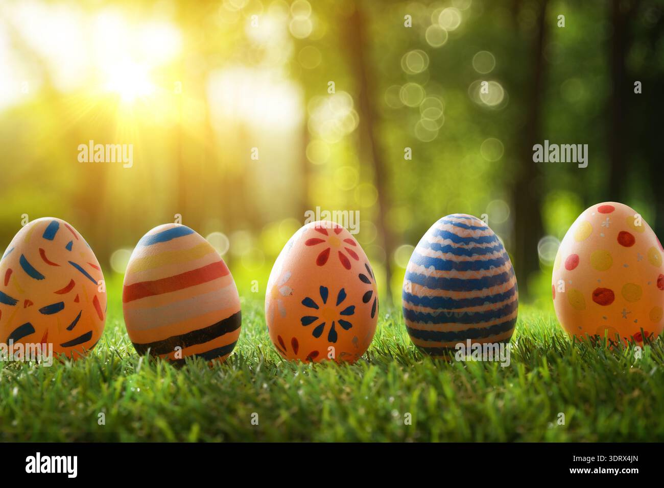 Fünf farbenfrohe Ostereier auf grünem Gras mit warmem Sonnenlicht und Waldbokeh Hintergrund, lebhafte Frühlingsfestszene. Stockfoto