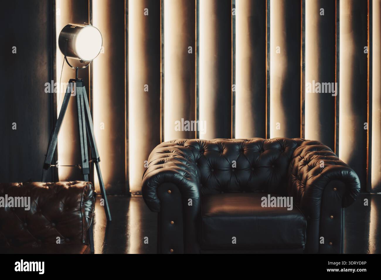 Luxuriöse Einrichtung mit schwarzem chesterfield-Ledersofa und -Sessel, Vintage-Studio-Spotlight auf Stativ, warme vertikale Wandleuchten, dunkles lou Stockfoto