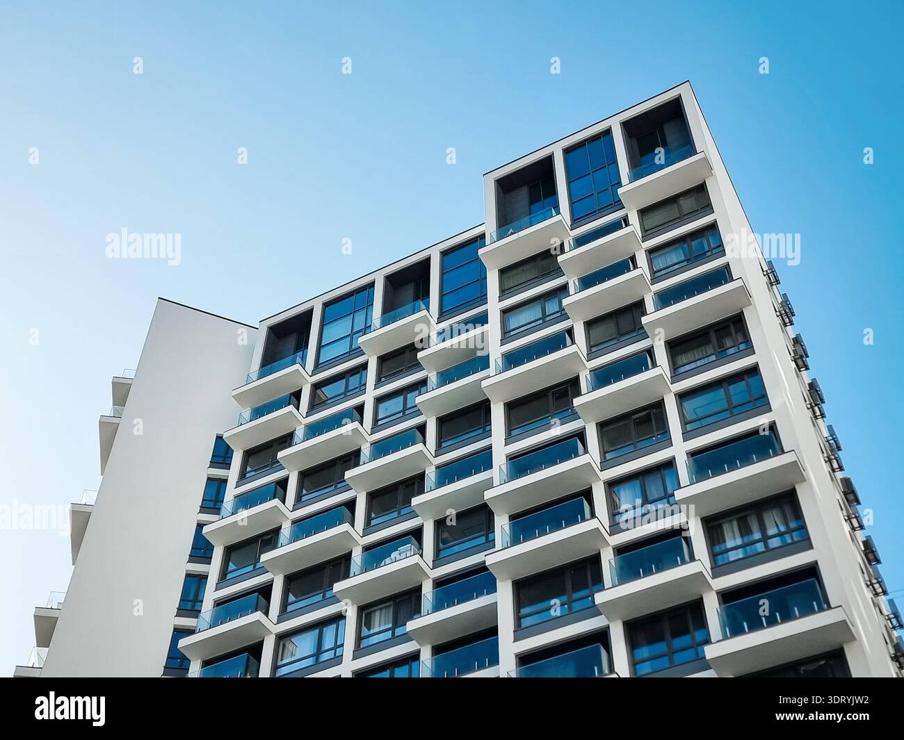 Modernes weißes Wohngebäude vor klarem blauem Himmel. Flacher Winkel von zeitgenössischen Hochhauswohnungen mit geometrischer Fassade und Glasbalkonen. Stockfoto