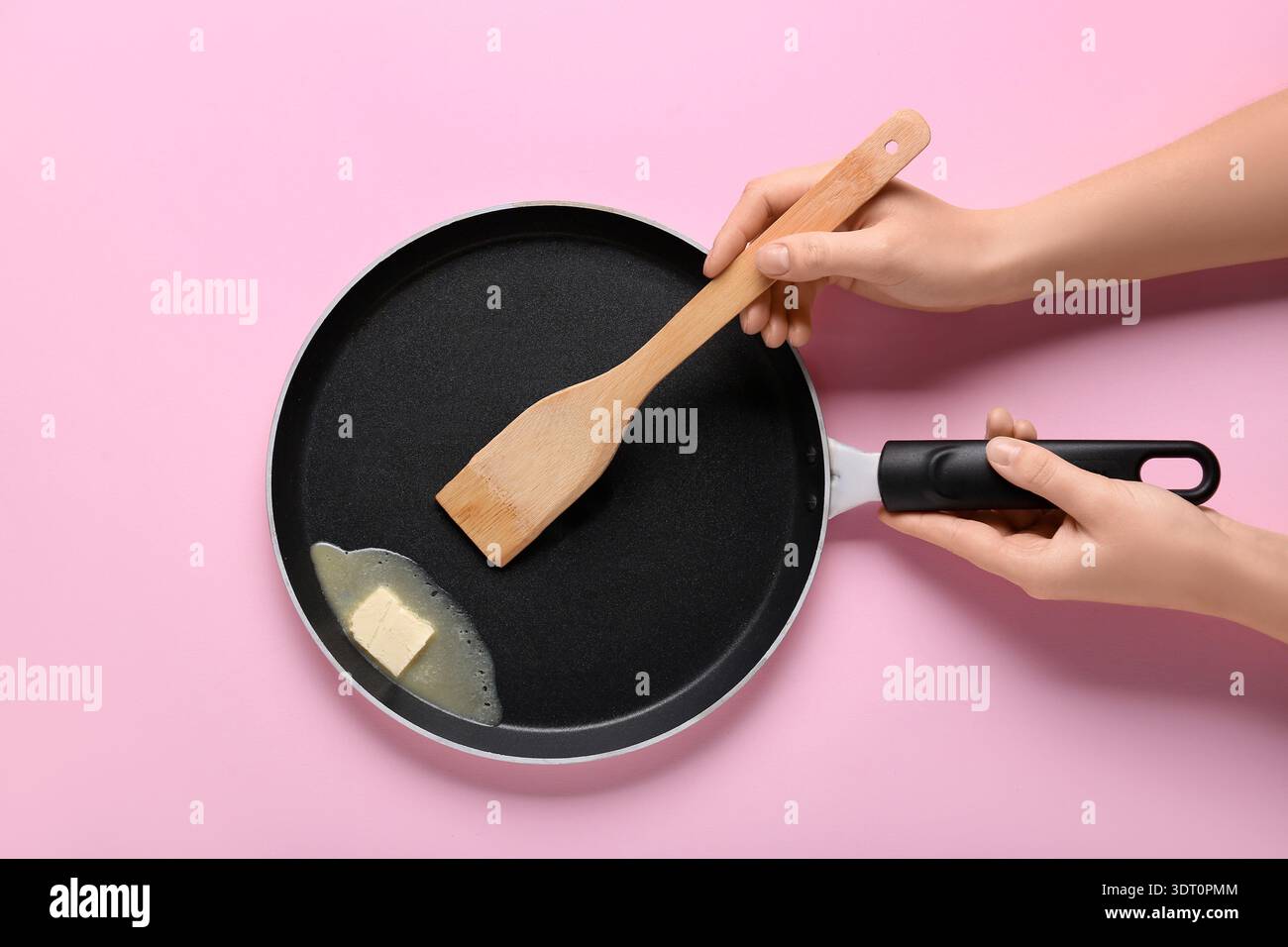 Weibliche Hände halten die Pfanne mit Butter und Spachtel auf rosafarbenem Hintergrund Stockfoto