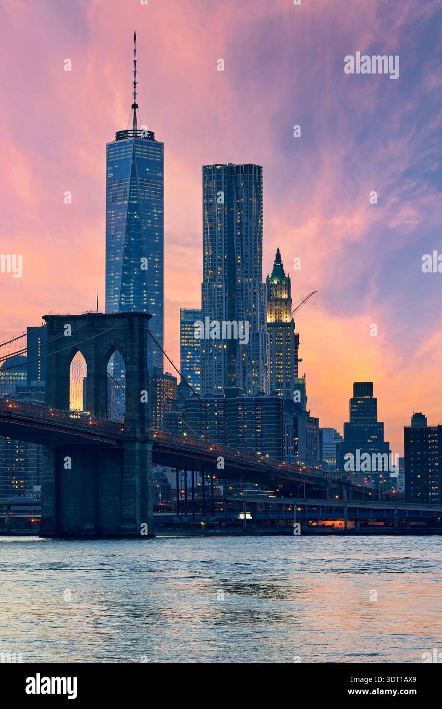 Die Brooklyn Bridge steht stark, wenn die Skyline von Manhattan bei Sonnenuntergang erleuchtet wird. Die Gebäude reflektieren Farben am Abendhimmel, während sich der Verkehr unter sich bewegt. Stockfoto
